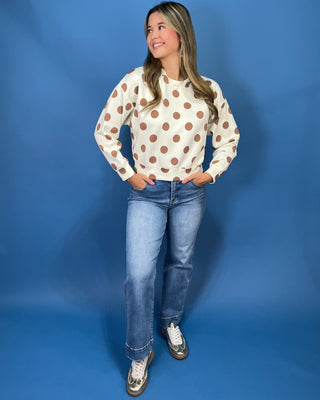 A woman modeling a cream long-sleeve crewneck sweatshirt with brown polka dots, paired with blue jeans and silver sneakers