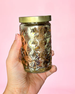 Hand holding a glass jar with a gold lid against a pink background