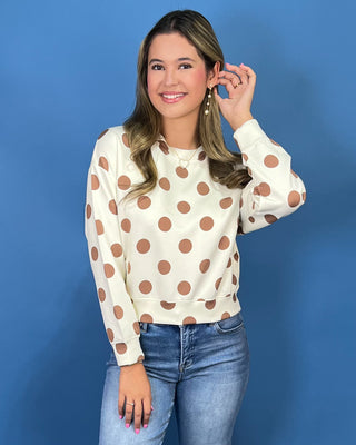 A woman modeling a cream long-sleeve top with brown polka dots, paired with blue jeans and metallic sneakers