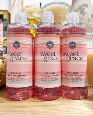 Three bottles of sweet grace dish soap on a wooden surface with a blurred background.