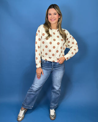 A woman posing in a cream long-sleeve sweatshirt with brown polka dots, blue jeans