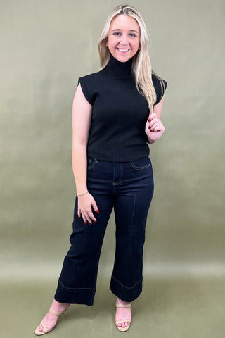 Woman wearing a black sleeveless top and dark jeans against a green background