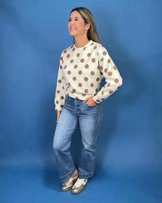 A woman modeling a cream long-sleeve top with brown polka dots, paired with blue jeans and metallic shoes