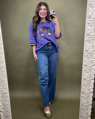 Woman taking a mirror selfie wearing a purple shirt with a graphic design and blue jeans on a green background.