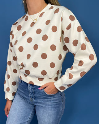 A woman wearing a cream-colored long-sleeve sweatshirt with large brown polka dots, paired with blue jeans, set against a solid blue background.