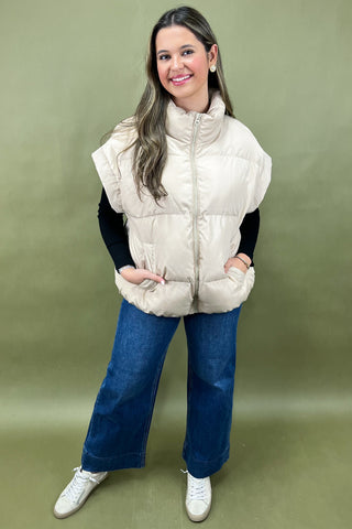 Woman wearing a beige puffer vest over a black long-sleeve shirt, blue jeans, and white sneakers on a green background.