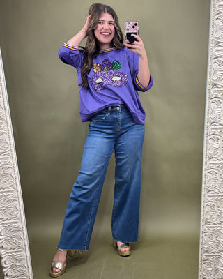Woman taking a mirror selfie wearing a purple sweater with sequin details and blue jeans on a green background.