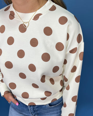 A person wearing a cream sweatshirt with large brown polka dots, paired with blue jeans and a gold heart necklace