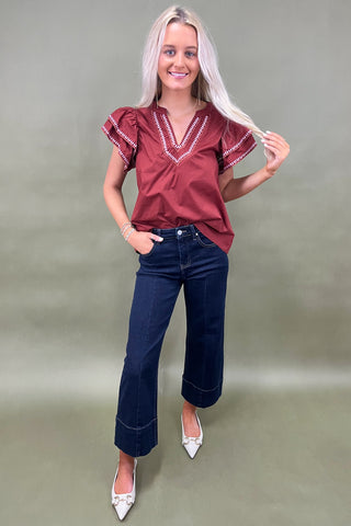 Woman wearing a burgundy blouse with ruffled sleeves and dark blue jeans on a plain background