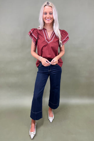 Woman wearing a burgundy blouse with ruffled sleeves and dark blue jeans on a plain background