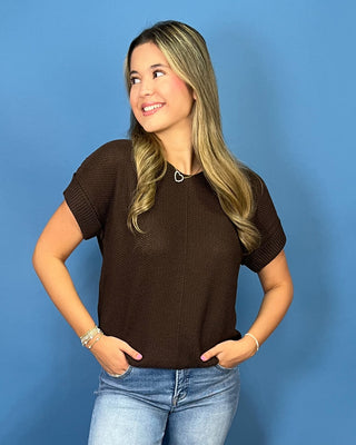 A woman with blonde-highlighted hair wearing a dark brown textured short-sleeve knit top and light-wash blue jeans, standing against a solid blue background.