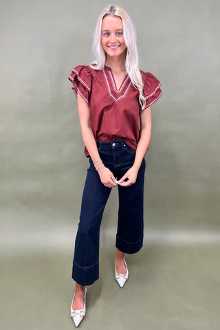 Woman wearing a burgundy blouse with ruffled sleeves and dark blue jeans on a plain background