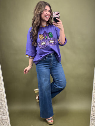 Woman taking a mirror selfie wearing a purple sweatshirt with a colorful design and blue jeans on a green background.