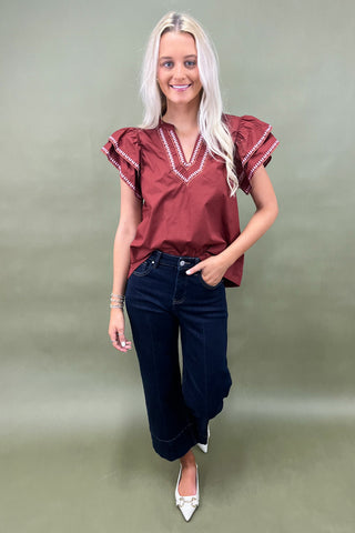 Woman wearing a burgundy blouse with ruffled sleeves and dark blue jeans on a plain background