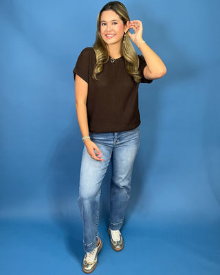 A woman modeling a dark brown short-sleeve top and light blue jeans against a solid blue studio backdrop.