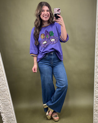 Woman taking a mirror selfie wearing a purple embellished top and blue jeans on a green background.