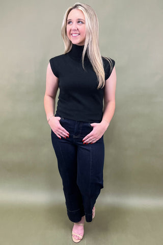 Woman wearing a black sleeveless top and dark jeans against a plain background