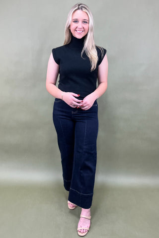 Woman wearing a black sleeveless top and dark jeans against a plain background