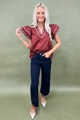 Woman wearing a burgundy blouse with ruffled sleeves and dark blue jeans on a plain background