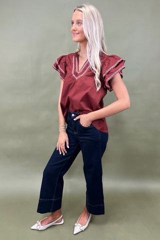 Woman wearing a burgundy blouse with ruffled sleeves and dark blue jeans on a plain background