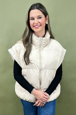Woman wearing a beige puffer vest over a black long-sleeve shirt on a green background