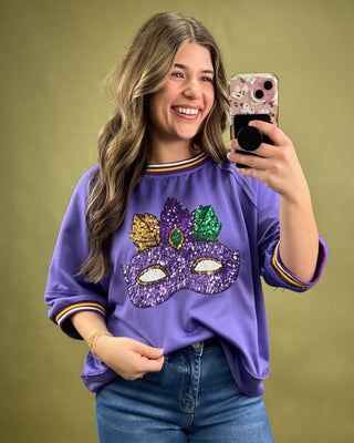 Woman taking a selfie wearing a purple sweatshirt with a mask design on a green background