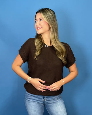 A woman with blonde-highlighted hair wearing a dark brown short-sleeve knit top and light blue jeans