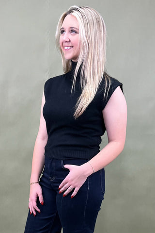 Woman wearing a black sleeveless top and dark jeans against a plain background