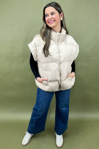 Woman wearing a beige puffer vest and blue jeans on a green background