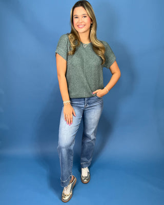 A woman modeling a short-sleeve knit top in a heathered sage green color, paired with light-wash blue jeans and metallic sneakers