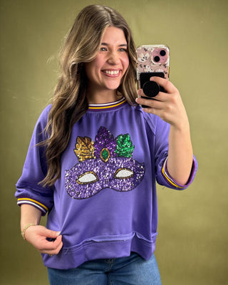 Woman taking a selfie wearing a purple sweatshirt with a mask design on a green background