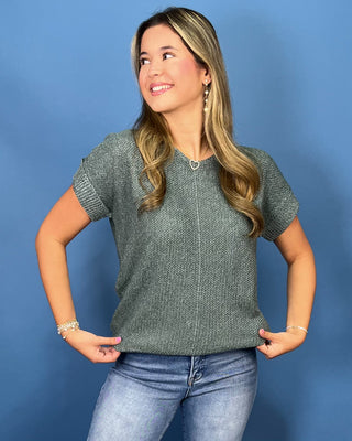 A woman wearing a textured gray short-sleeve knit top and blue jeans, posing against a solid blue studio background.
