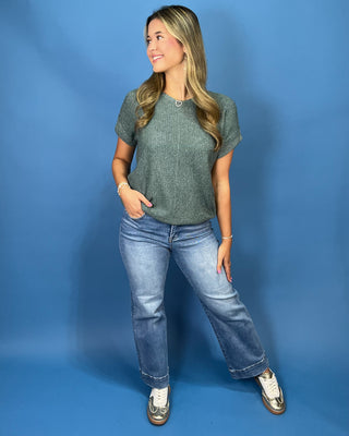 A woman modeling a short-sleeve textured knit top in sage green, paired with light-wash blue jeans and metallic sneakers, standing against a solid blue studio backdrop.