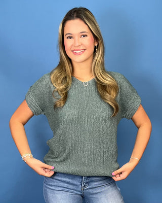 A woman with blonde hair wearing a textured sage green short-sleeve knit top and blue jeans, posing against a solid blue background.