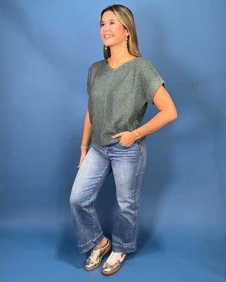 A woman modeling a short-sleeve textured sage green top and light-wash blue jeans against a solid blue studio backdrop.