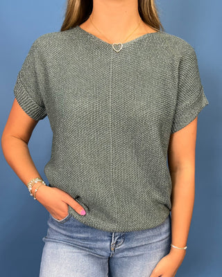 A woman wearing a short-sleeve knit top in a heathered gray-green color, paired with light blue jeans, standing against a solid blue background.