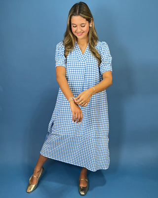 A woman modeling a short-sleeve blue and white gingham midi dress with a tiered skirt, standing against a solid blue studio backdrop.