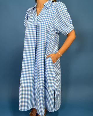 A woman wearing a light blue and white gingham midi dress with short puff sleeves, a collar, and side pockets, standing against a solid blue background.