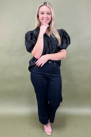 Woman wearing a black top and dark jeans against a green background