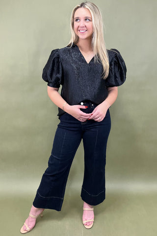 Woman wearing a black blouse with puffed sleeves and dark pants against a green background