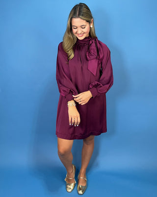 A woman modeling a long-sleeve burgundy dress with a high neck and large bow detail at the collar, standing against a solid blue studio backdrop.