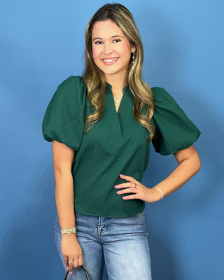 A woman modeling a dark green short-sleeve top with puffed sleeves and a split V-neckline, paired with blue jeans