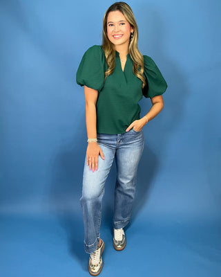 A woman modeling a dark green textured puff-sleeve top with a split neckline, paired with light-wash blue jeans and metallic sneaker