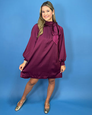 A woman modeling a long-sleeve, high-neck, satin-like mini dress in deep burgundy with a large bow detail at the neck, standing against a solid blue studio backdrop.