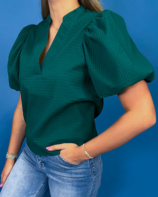 A woman wearing a dark green textured puff-sleeve blouse with a split neckline, paired with light blue jeans