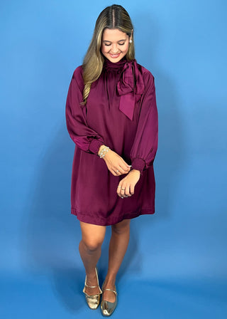 A woman modeling a long-sleeve, high-neck mini dress in deep burgundy with a large bow detail at the neck, standing against a solid blue studio backdrop.