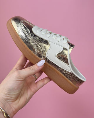 Gold and white sneaker with brown sole held by a hand against a pink background