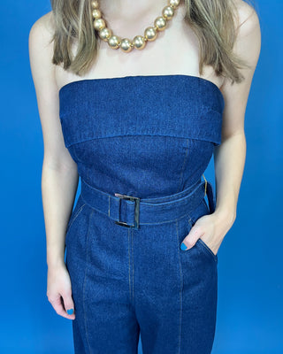 A person wearing a blue denim strapless jumpsuit with a matching belt and silver buckle, styled with a gold beaded necklace