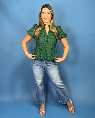 A woman modeling a dark green eyelet lace peplum top with puff sleeves, paired with light-wash blue jeans and beige sandals, standing against a solid blue studio backdrop.