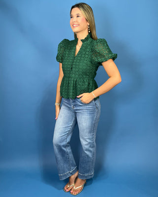 A woman modeling a dark green eyelet lace blouse with puff sleeves and a peplum waist, paired with light-wash blue jeans
